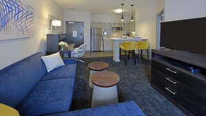 A modern living room with a blue sofa, two round wooden side tables, a flat-screen TV, and a kitchen area with a dining table and yellow chairs. - Free Breakfast. Gym. Near Medical Center. (Little Rock)