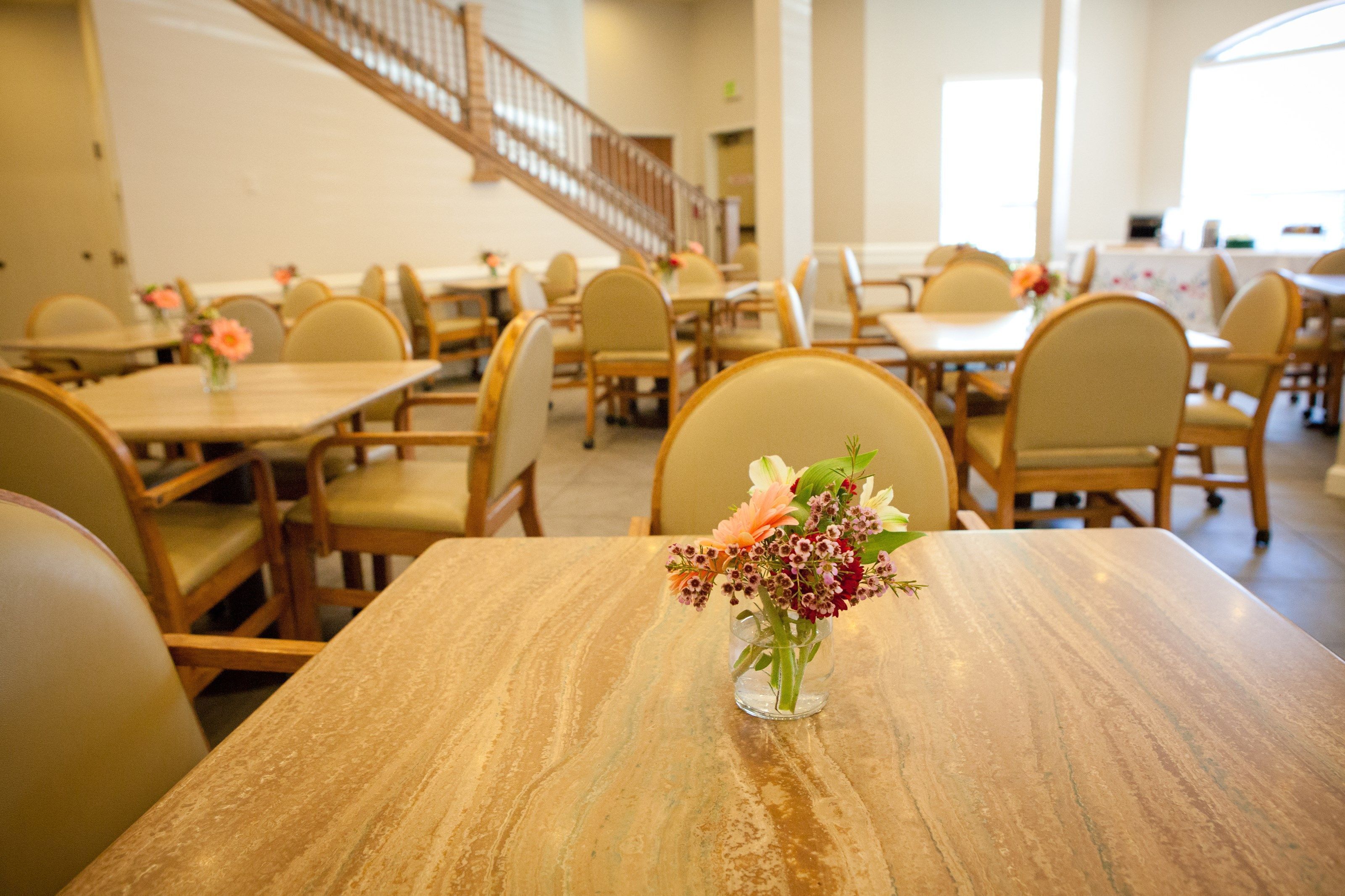 A dining area with wooden tables and chairs, a staircase in the background, and a vase with flowers on one table.