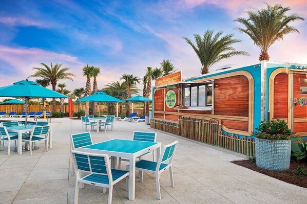 An outdoor dining area with tables and chairs, a food truck, and palm trees.