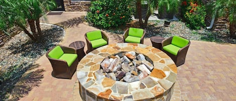 Un patio exterior con un hogar a leña de piedra rodeado de sillas de mimbre con cojines verdes.