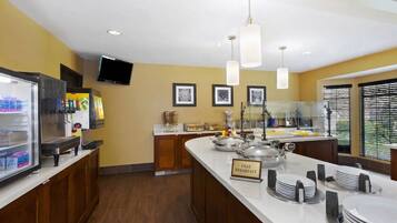 A breakfast bar with a sign indicating free breakfast, a refrigerator with beverages, and a television mounted on the wall.