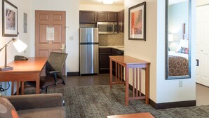 Habitación de hotel compacta con kitchenette, comedor con mesa y sillas, un pequeño sofá y una cama con cabecera estampada.