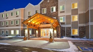 Un hotel de varios pisos con fachada de piedra, un acceso cubierto y un estacionamiento con nieve.