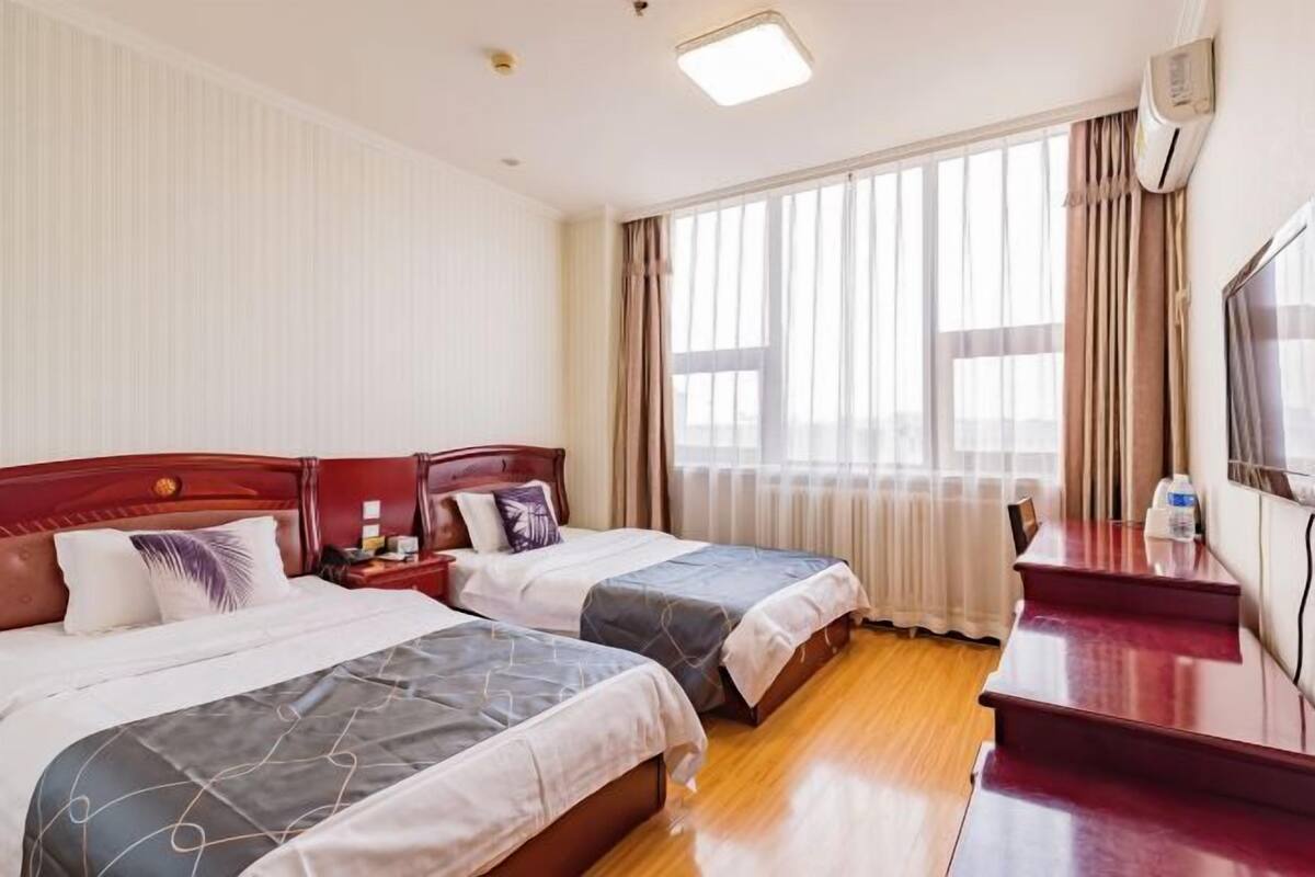 A hotel room with two beds, a desk, a TV, and a large window with curtains.