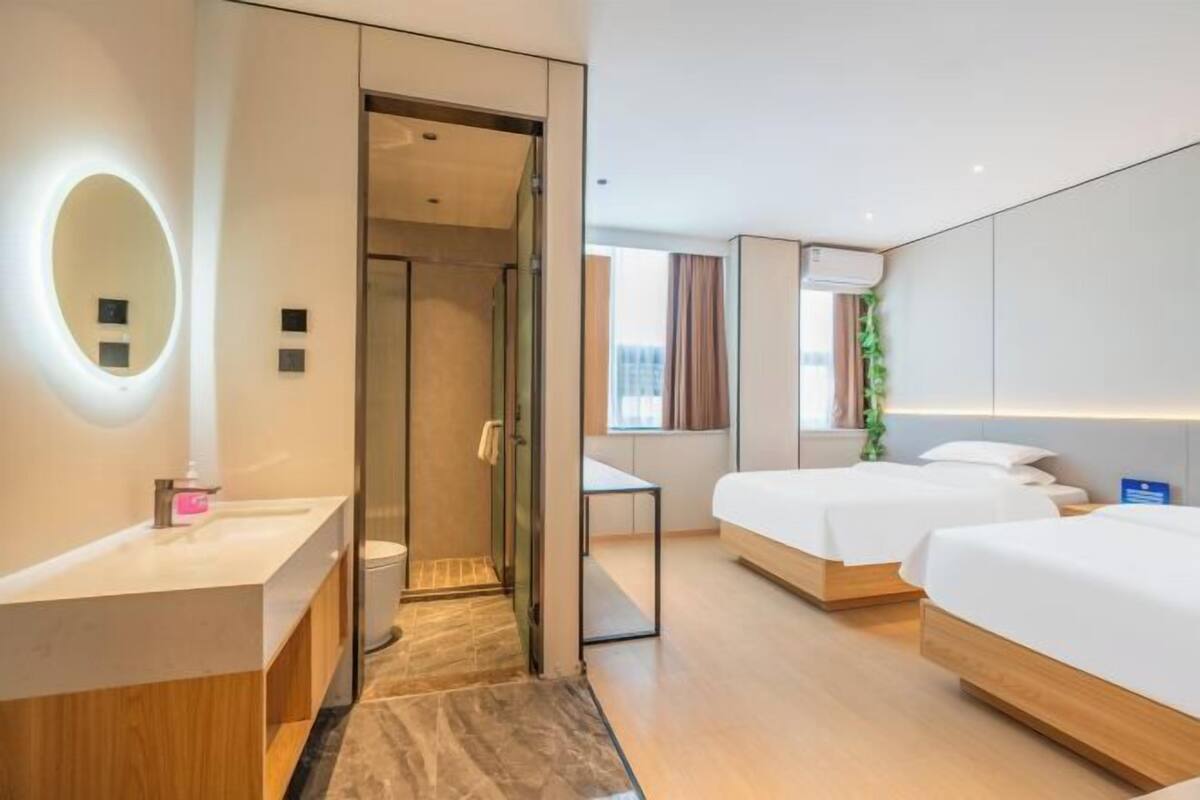 A hotel room with a modern design, featuring a large mirror, a wooden vanity, two beds, a small table, and a view of the city.