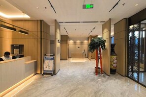 Un hall d’hôtel moderne avec un comptoir d’accueil, un panneau d’affichage numérique et une grande plante en pot.