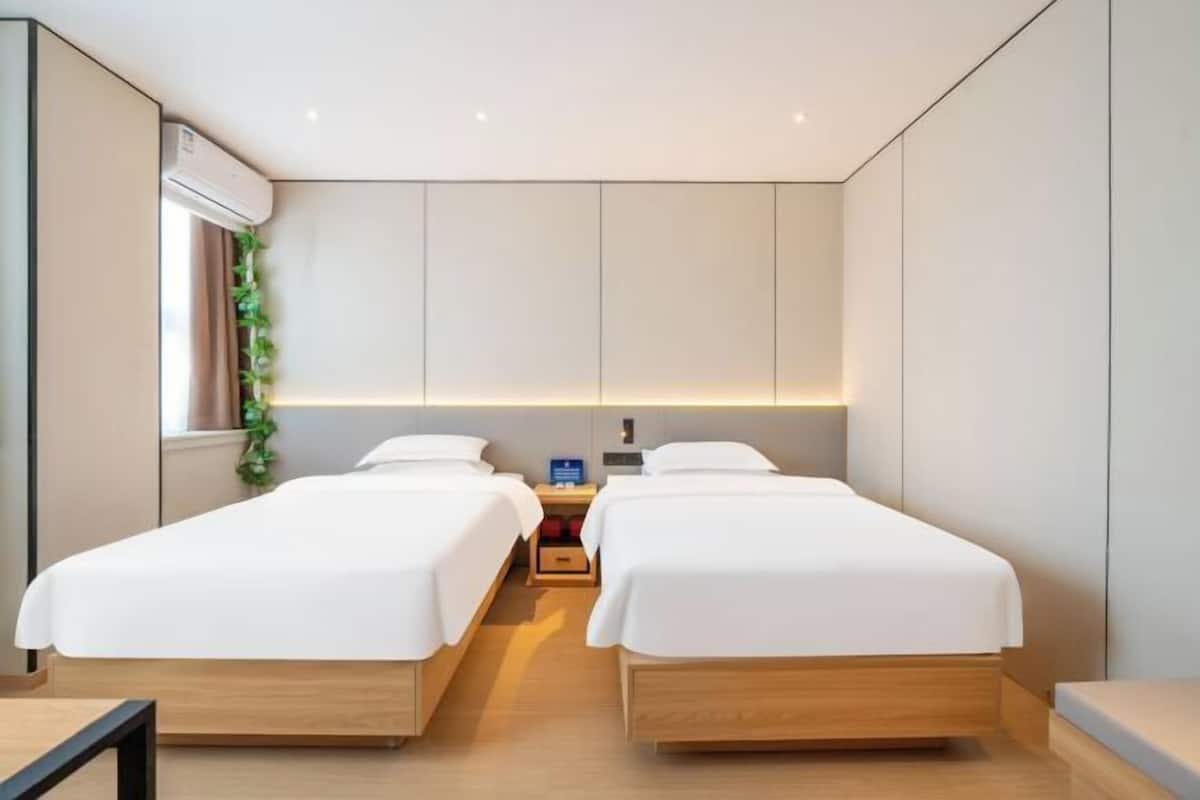 A modern hotel room with two single beds, a bedside table, a nightstand, and a window with curtains.