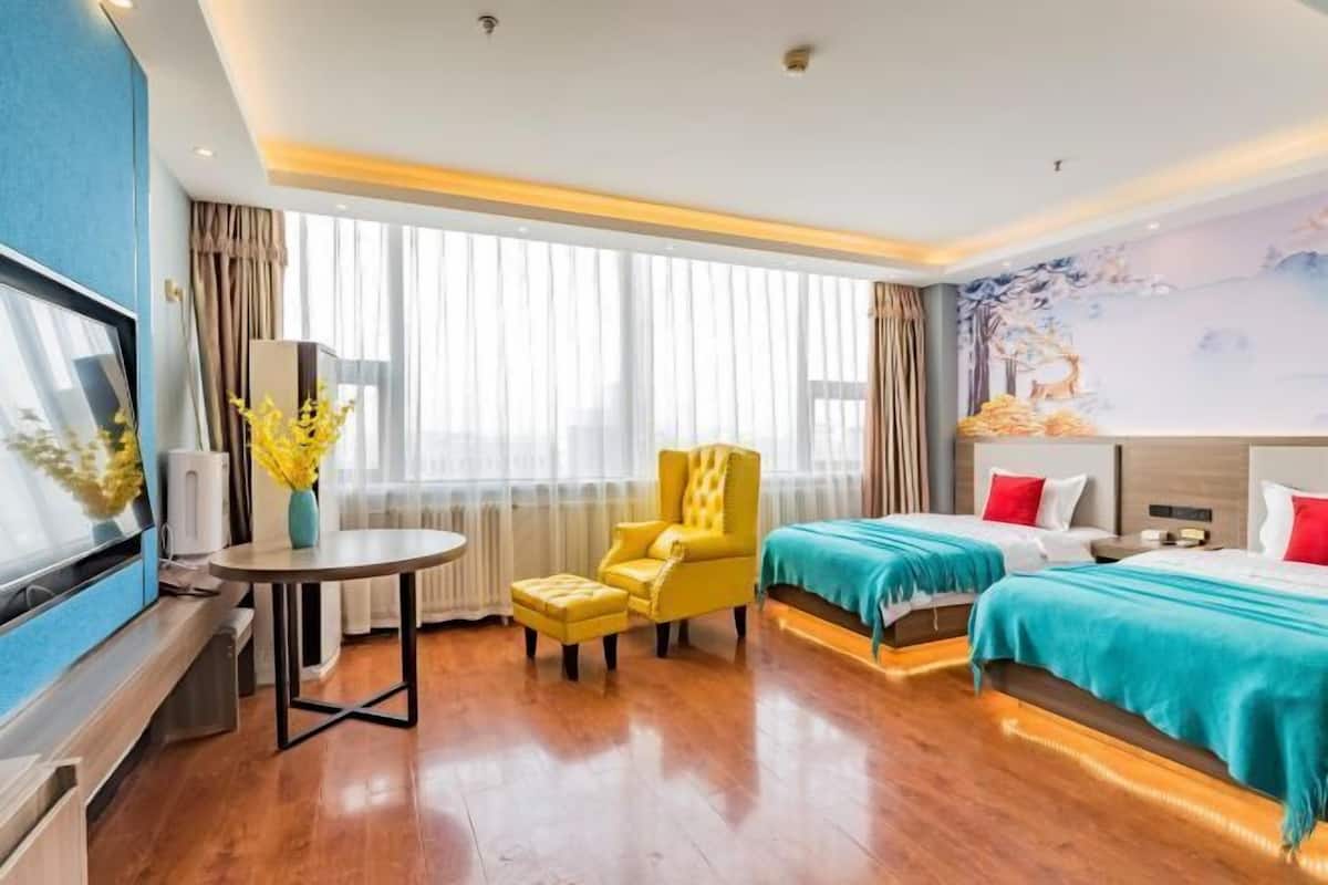 A hotel room with a wooden floor, two beds with teal and red bedding, a yellow armchair, a round table with a vase of yellow flowers, a TV, and a large window with sheer curtains.