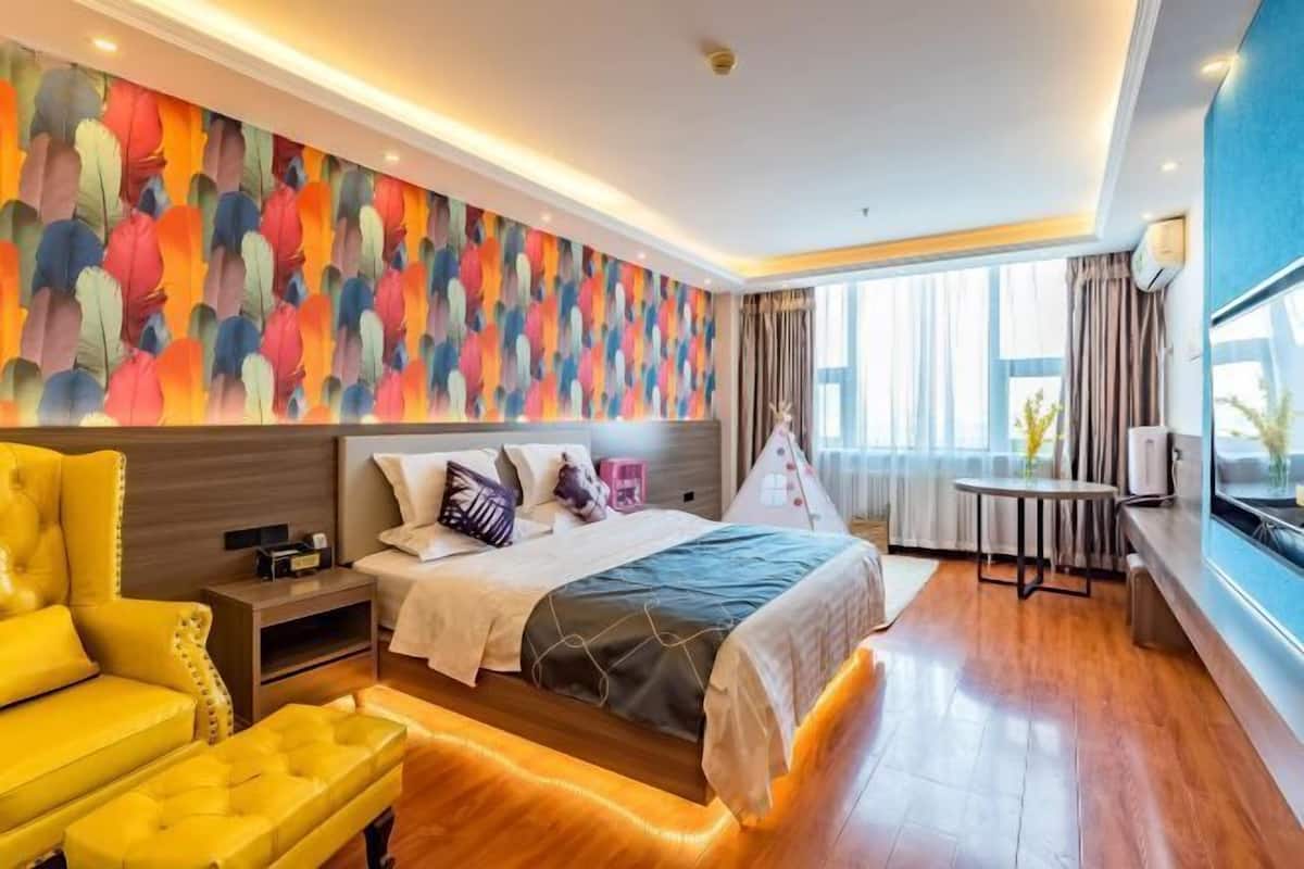 A modern hotel room with a large bed, a yellow armchair, a small table, and a colorful abstract wall mural.