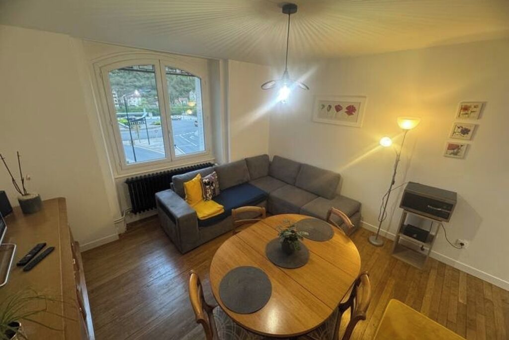 Un salon moderne avec une table à manger en bois, un canapé d’angle gris et une grande fenêtre donnant sur la rue.