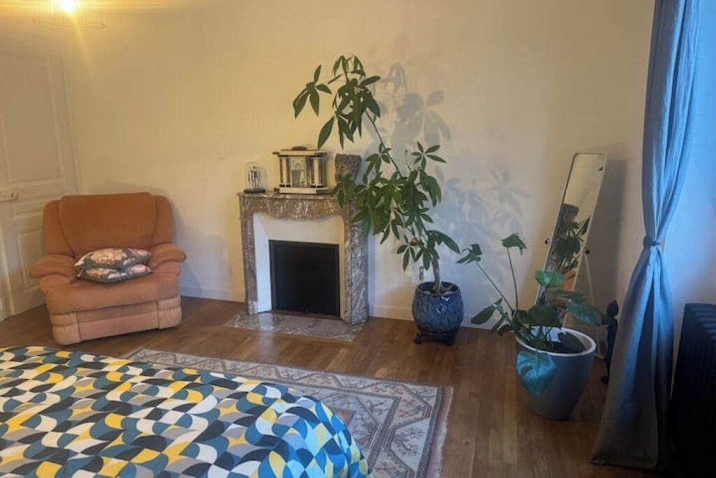 A bedroom with a patterned bedspread, a chair with a cushion, a fireplace, and two potted plants.
