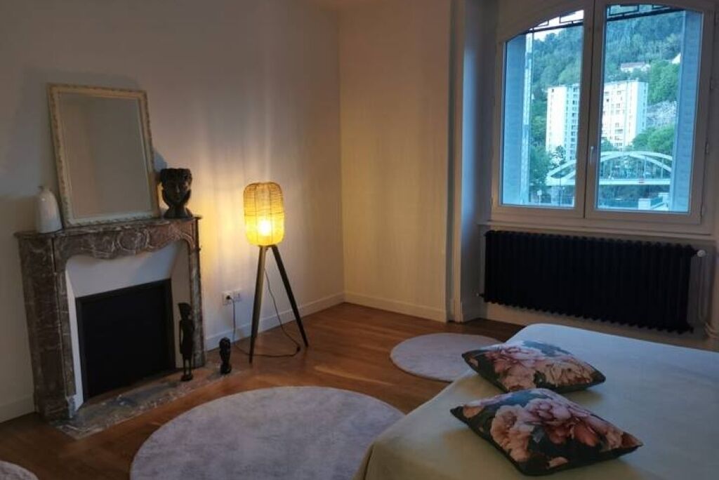 A room with a fireplace, a standing lamp, a bed with floral pillows, and a window with a view of buildings and greenery.