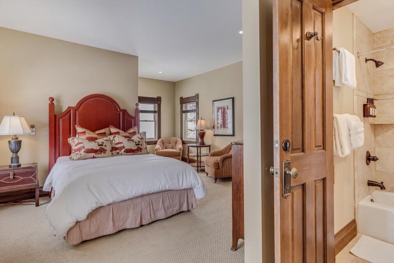 A hotel room with a large bed, a wooden door, a bathroom with a bathtub, and a chair by the window.
