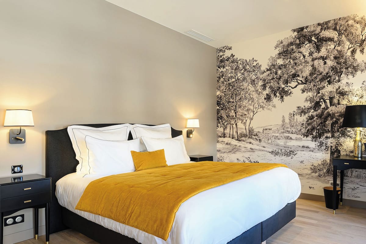 A modern bedroom with a large bed, two bedside tables, a wall-mounted lamp, and a mural of trees.