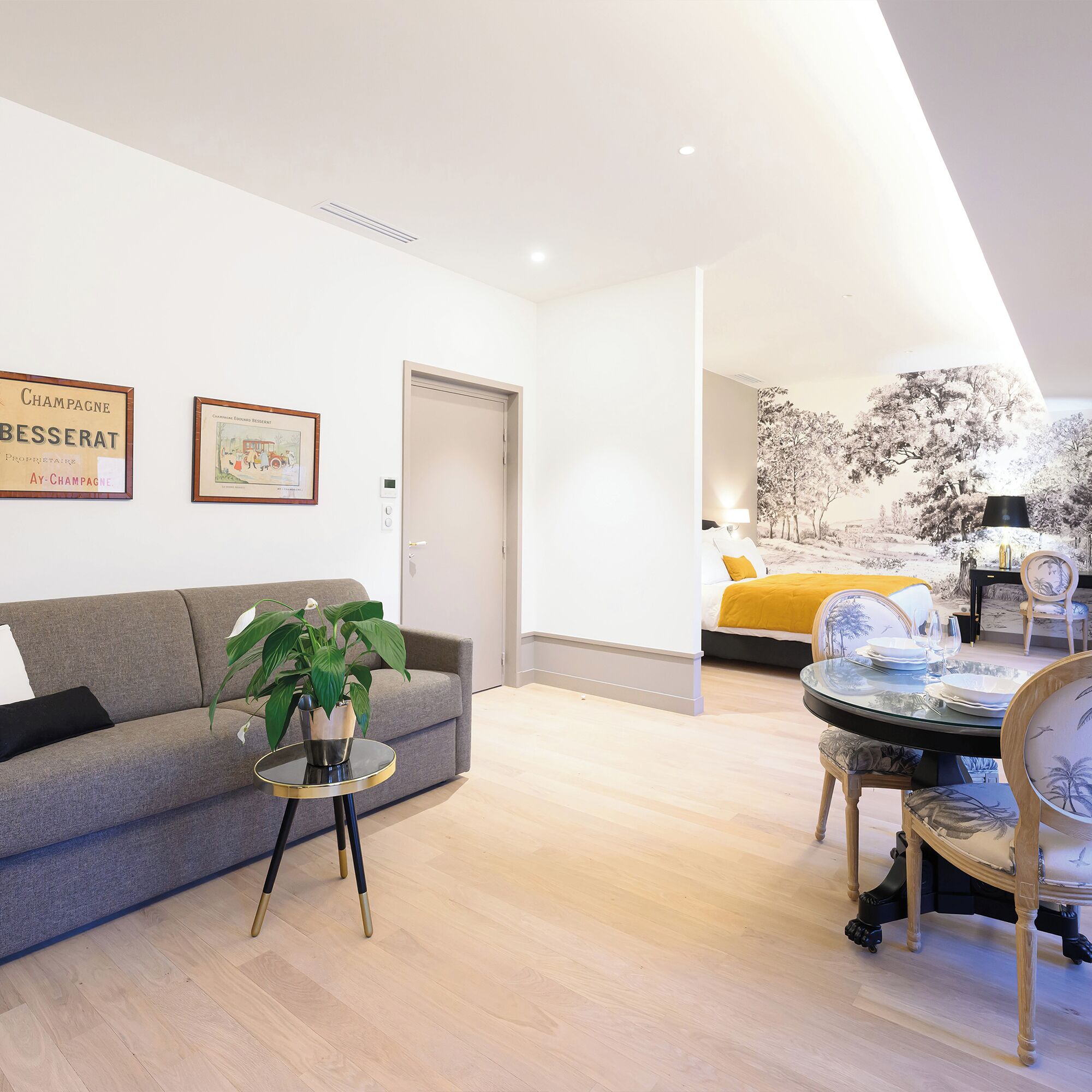 A modern living room with a grey sofa, a small round table with a plant, and a dining area with a glass table and chairs.
