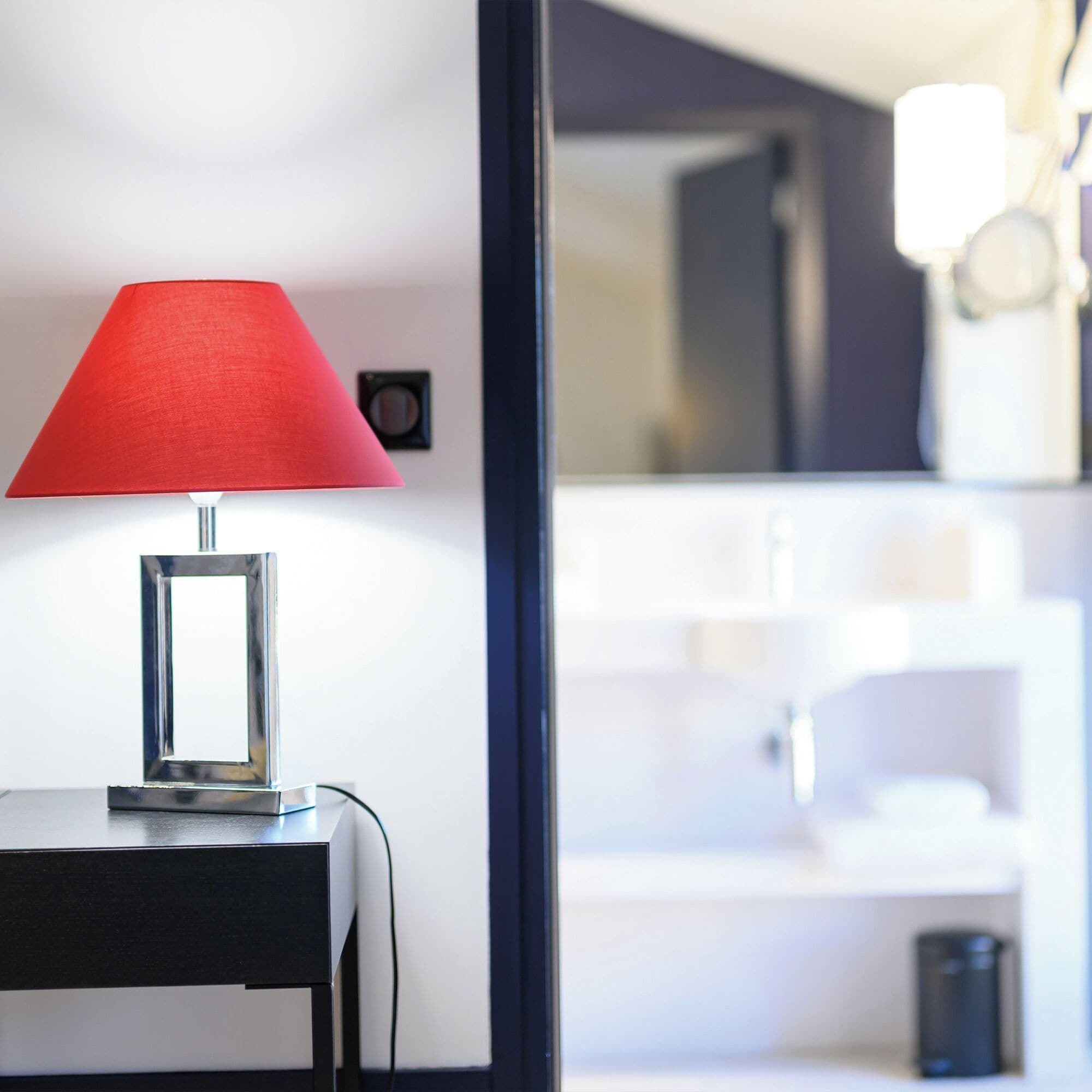 A modern hotel room with a red lamp on a bedside table, a mirror, and a view of the bathroom.