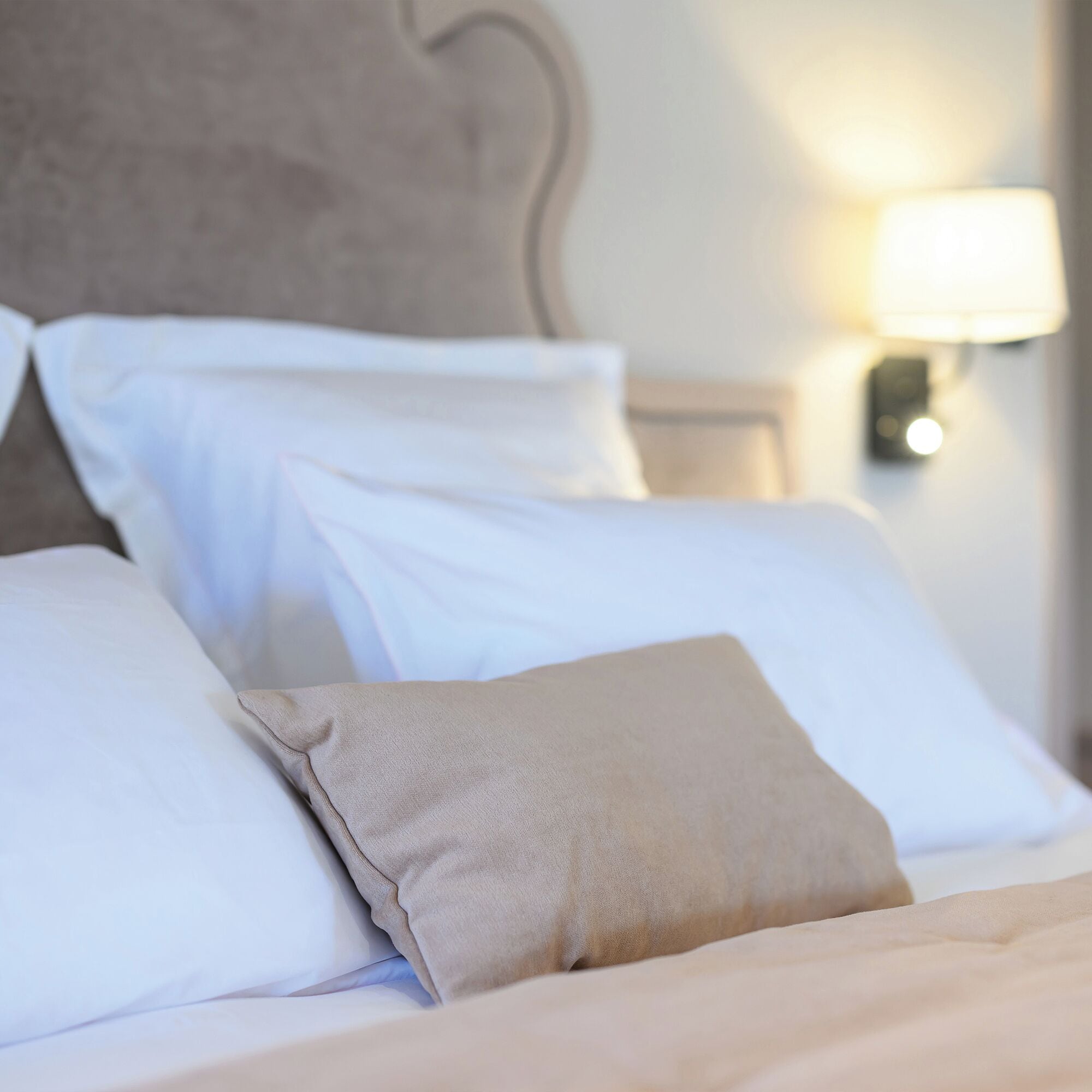 A neatly made bed with white and beige pillows, a headboard, and a wall-mounted lamp.