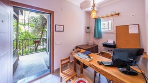 Ein Zimmer mit Schreibtisch aus Holz, einem Computer, einem Stuhl, einem kleinen Tisch, einem Fenster mit Vorhang und einer Tür, die auf einen Balkon führt.