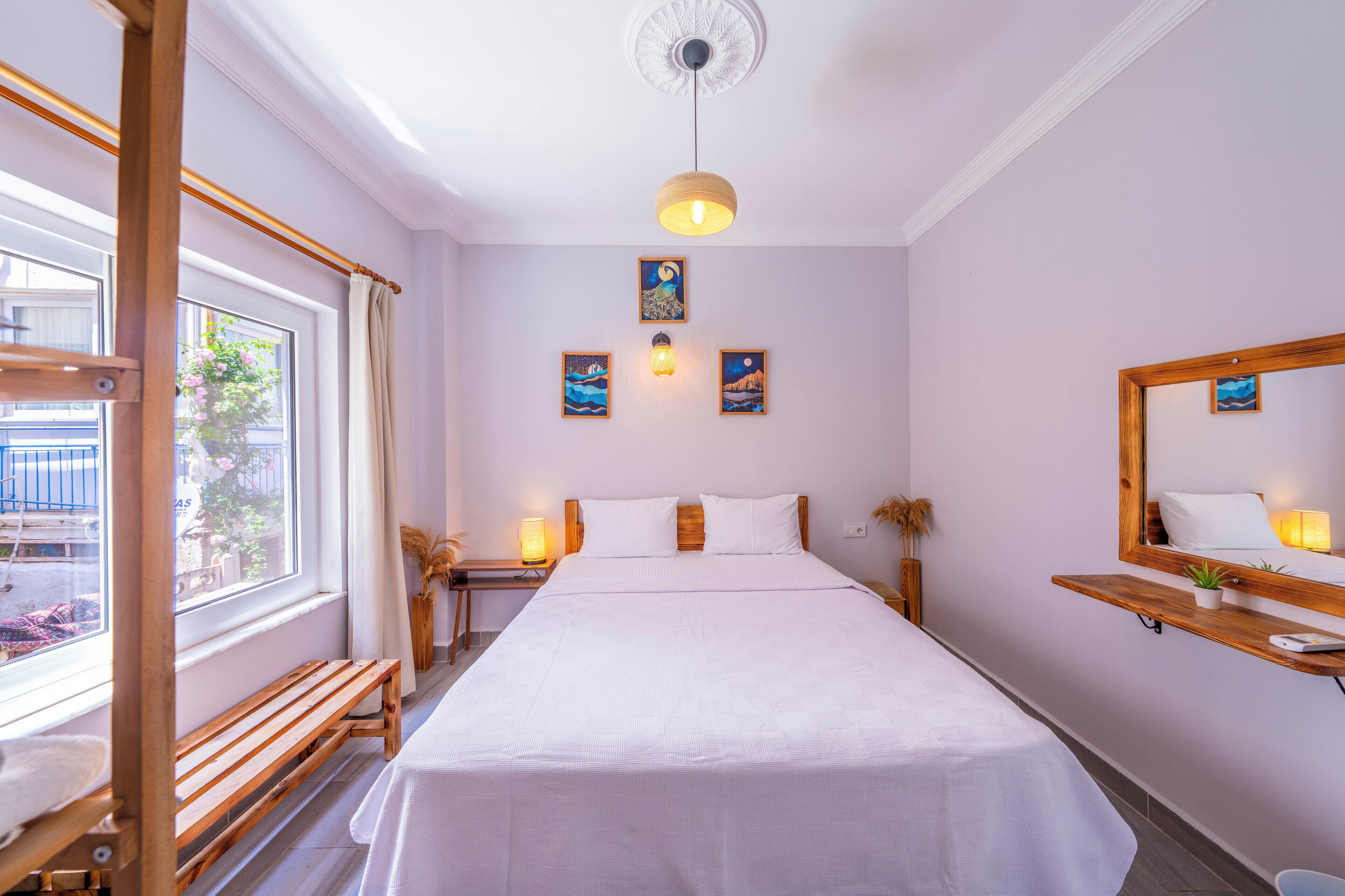 A neatly arranged bedroom with a large bed, bedside tables with lamps, a wooden bench, a window with curtains, and wall-mounted artwork.
