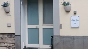 Une entrée d’immeuble avec une porte en verre dépoli, une enseigne indiquant « San De Bell & Breakfast », et deux plantes en pot de chaque côté.