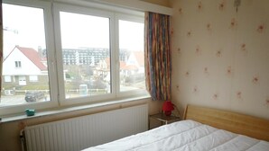 A bedroom with a bed, a window with curtains, a radiator, and a view of buildings outside. - Well-kept Corner Apartment in Koksijde (Koksijde)