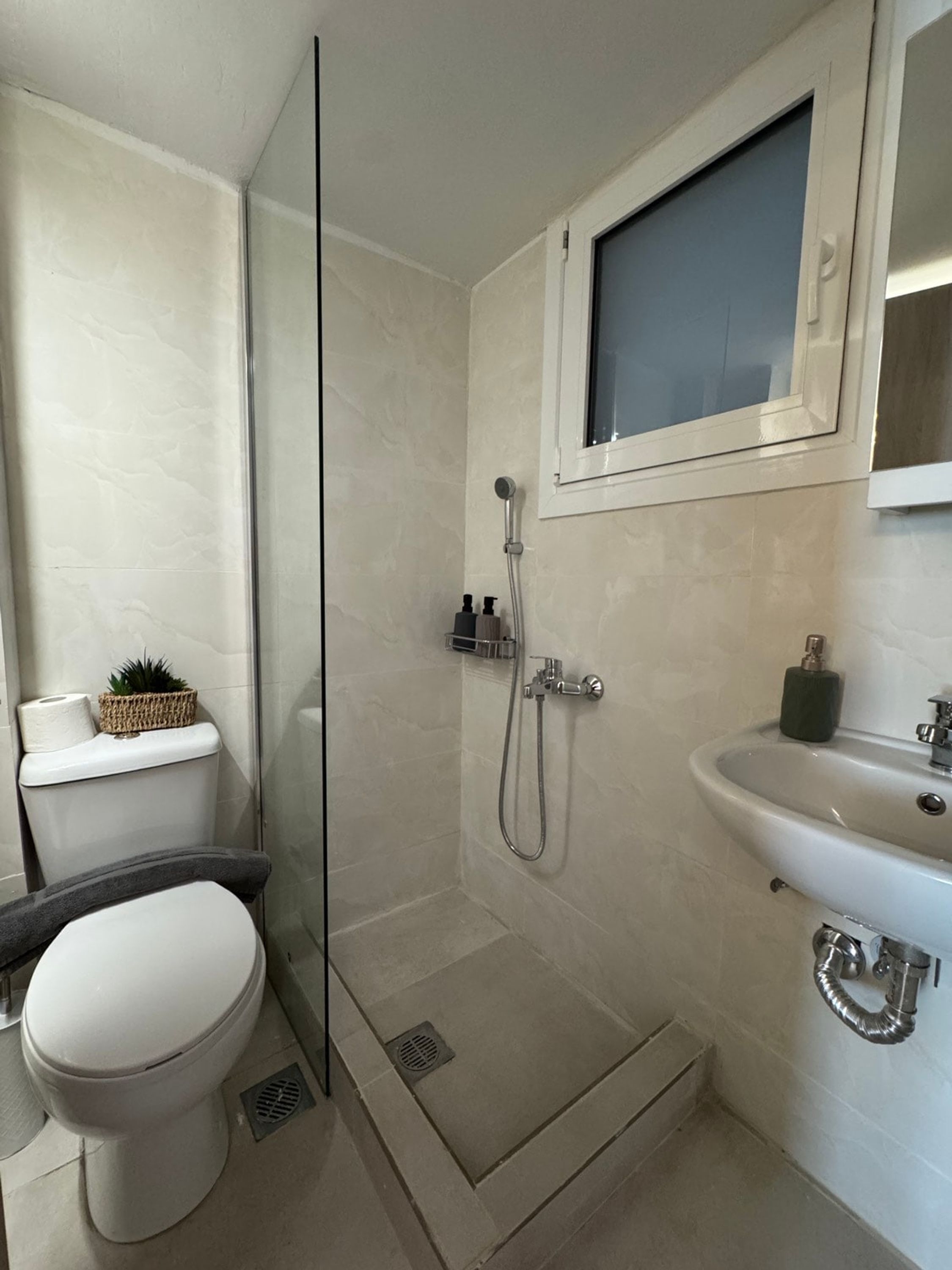 Een moderne badkamer met een douchecabine van glas, een toilet, een wastafel en een klein raam boven de spiegel.