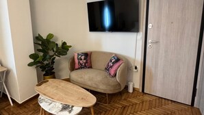 A room with a herringbone wooden floor, a beige sofa with colorful cushions, a small round table, a potted plant, and a flat-screen TV mounted on the wall. - Athenian Stay Chara s 2 Apartment (Chalandri)