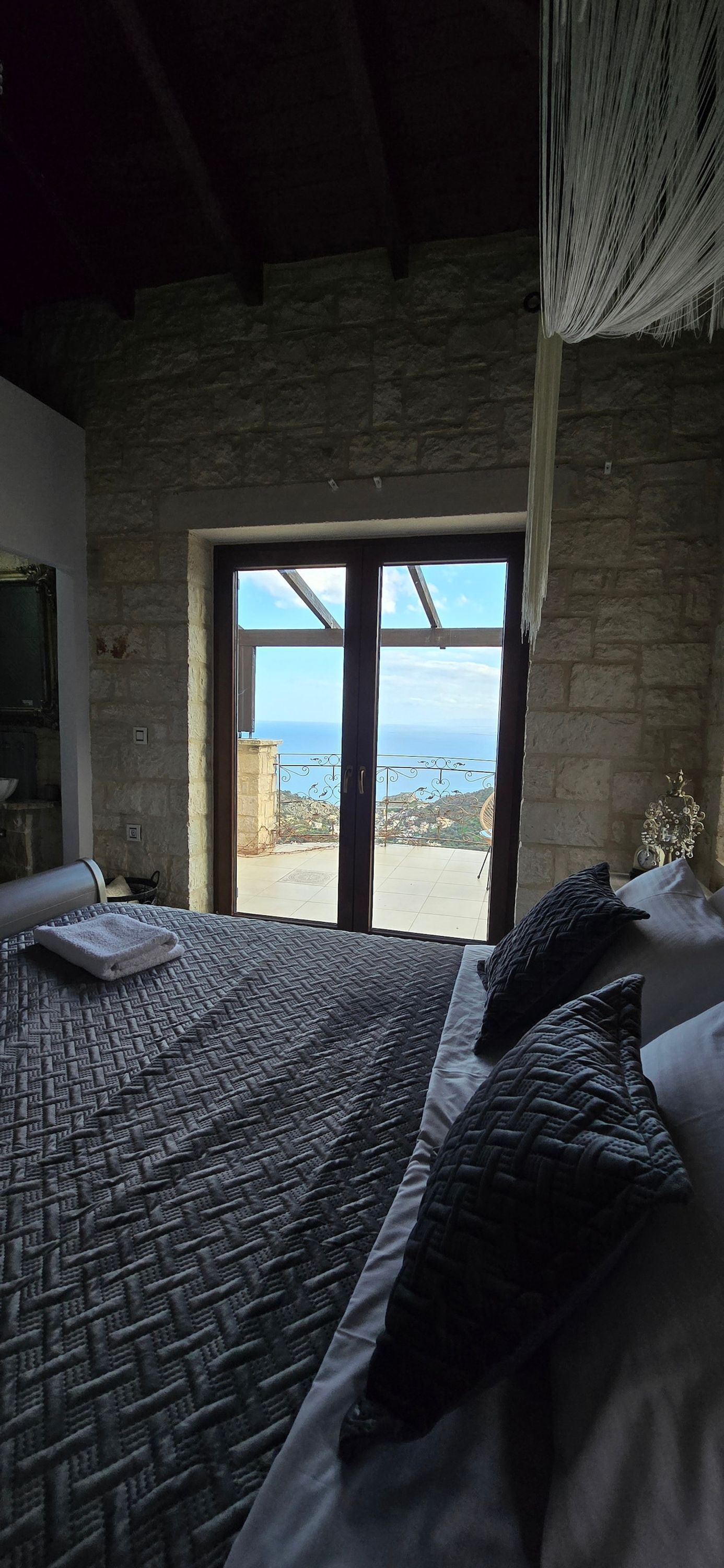 A bedroom with a large bed, a stone wall, and a view of the sea through a glass door.