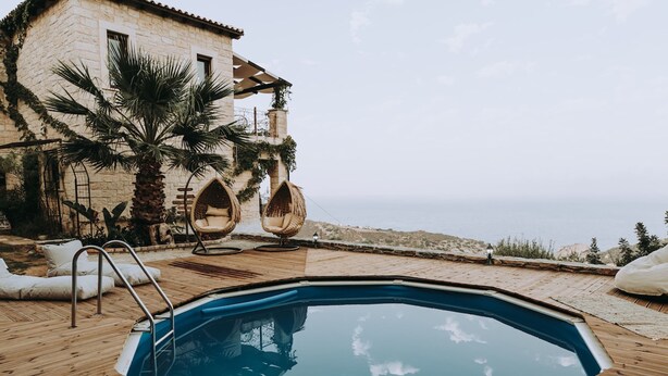 A swimming pool with wooden deck, lounge chairs, and a stone building with a view of the sea. - Luxury Villa Rodianna (Malevizi)