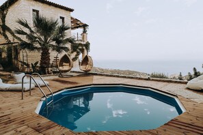 Piscina con muelle de madera, sillones de descanso y un edificio de piedra con vistas al mar.