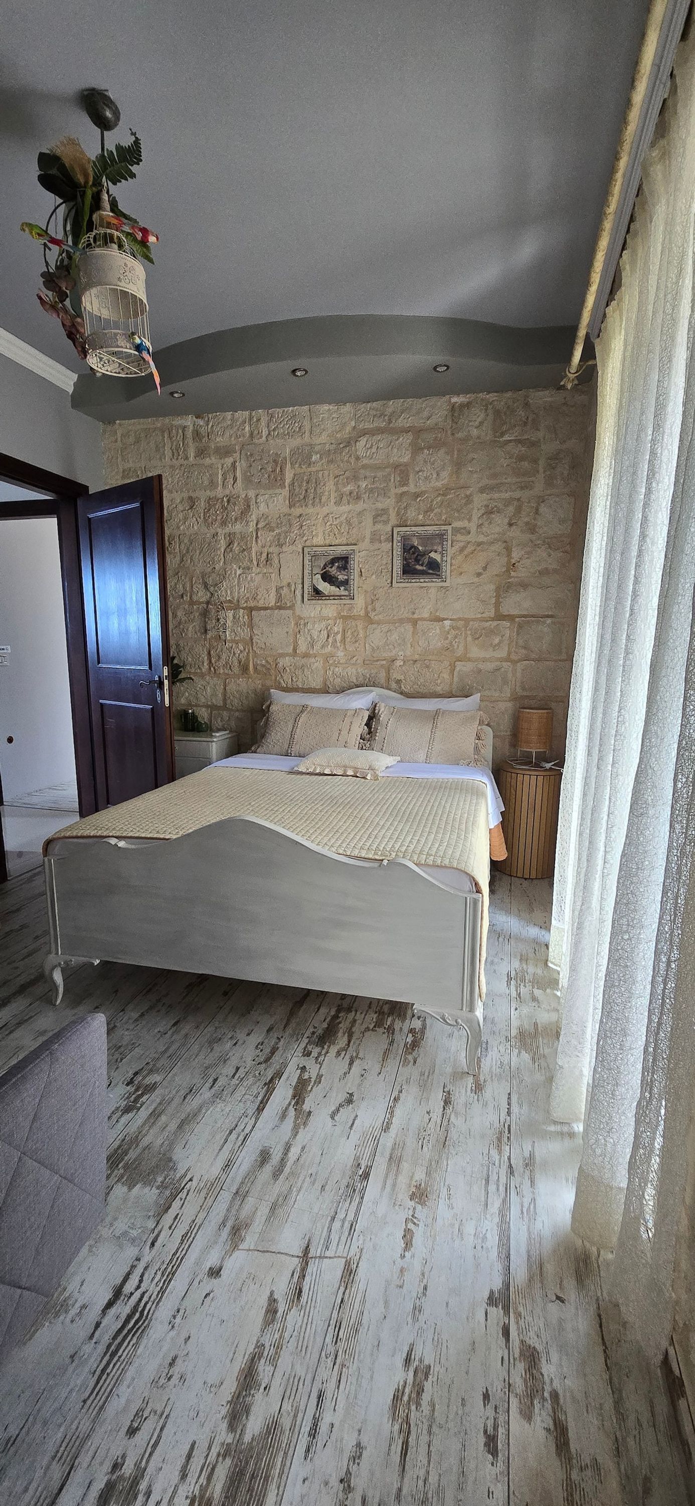 A bedroom with a stone wall, a bed with a white headboard, a nightstand, and a hanging plant.
