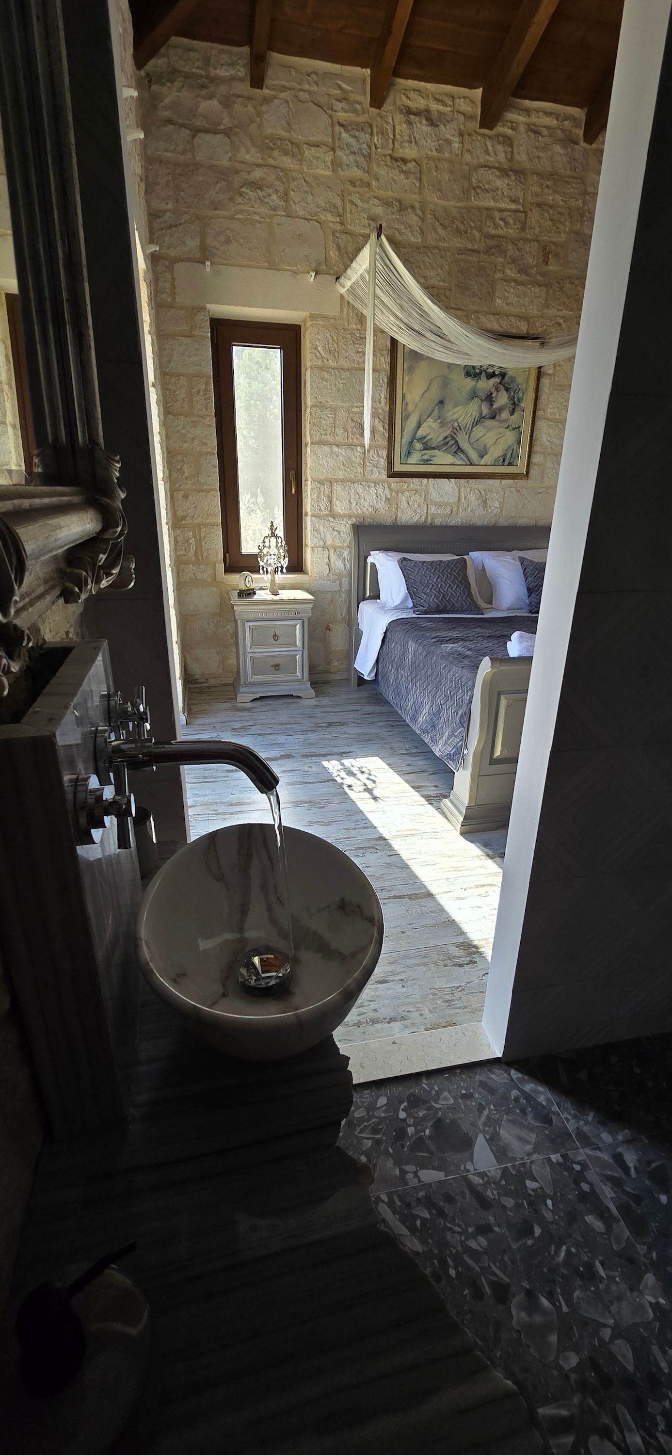 A stone-walled bedroom with a bed, a nightstand, a window, and a chandelier.