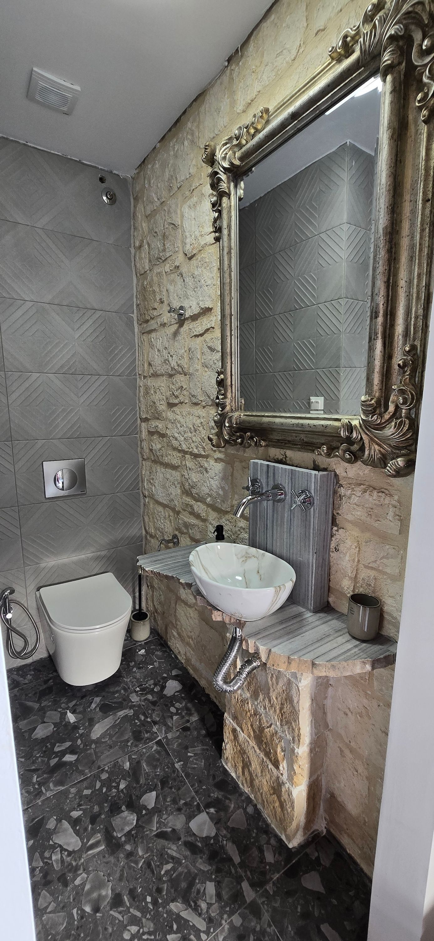 Un bany amb paret de pedra, un vàter blanc, un lavabo de marbre i un mirall gran i ornamentat.
