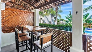 An outdoor patio with wooden tables and chairs, a wooden lattice wall, and a view of a pool and palm trees. - Modern Split-Level 2-Bedroom Condo with Pools, Terrace & Easy Beach Access (Sardinal)