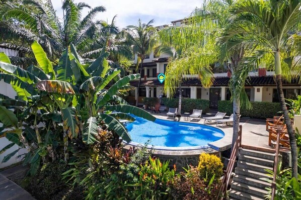 A swimming pool surrounded by lush tropical plants, with a building in the background. - Modern Split-Level 2-Bedroom Condo with Pools, Terrace & Easy Beach Access (Sardinal)