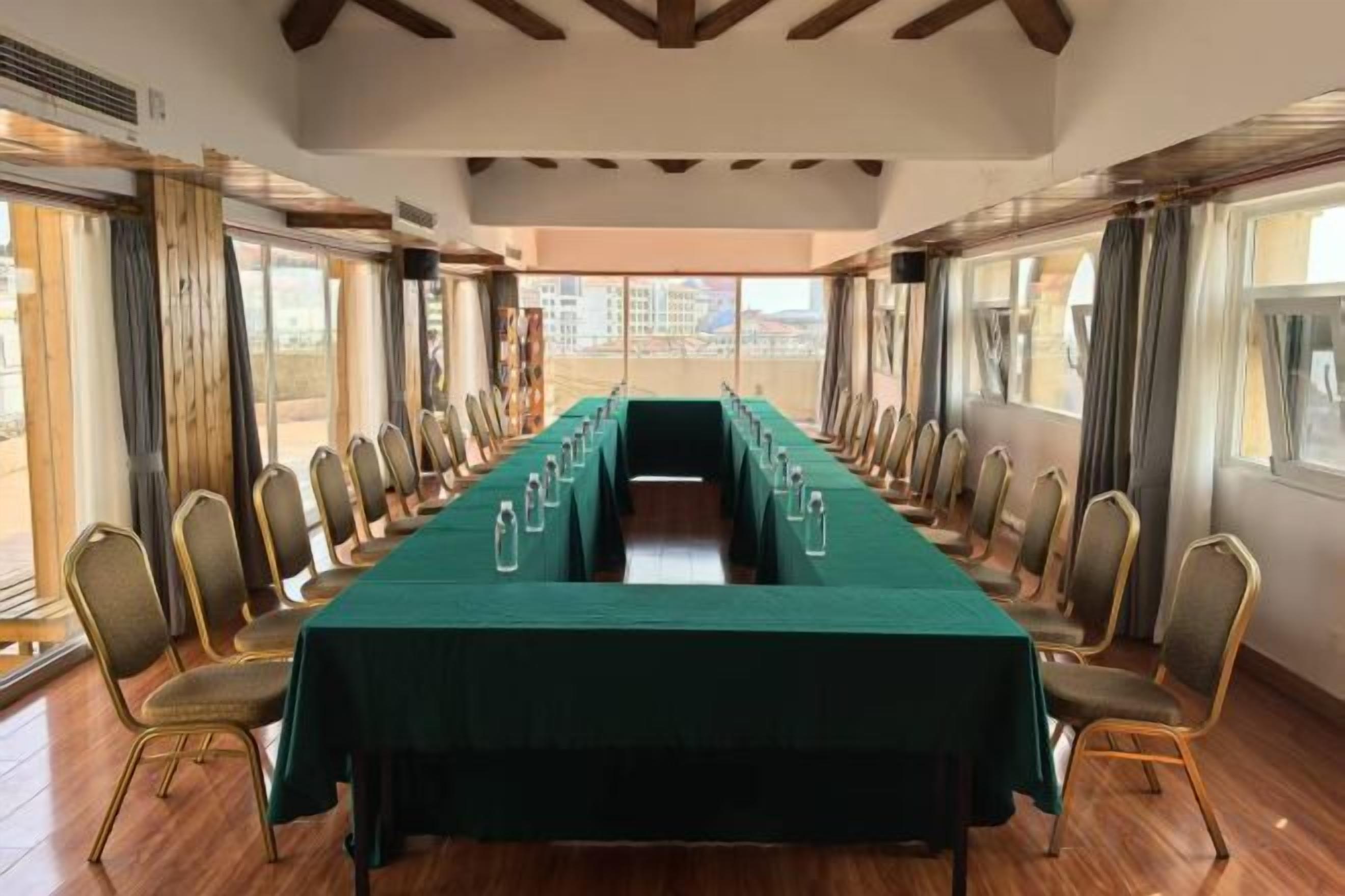 Un salón de conferencias con una larga mesa y sillas, ventanas grandes y vistas a edificios.