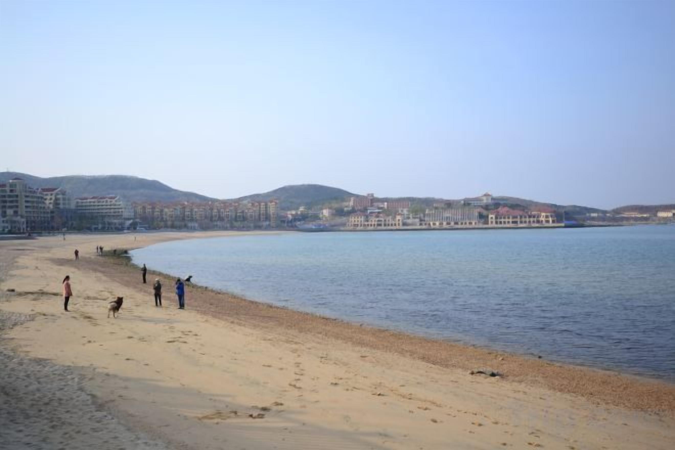 A beach with people walking along the shore, a calm sea, and buildings in the background.