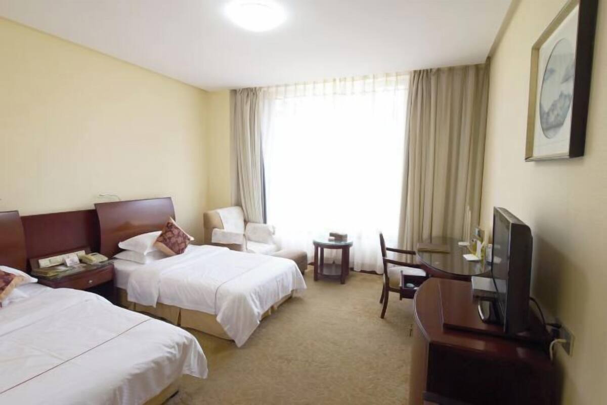 A hotel room with two beds, a desk with a TV, a chair, a small table, and a window with curtains.
