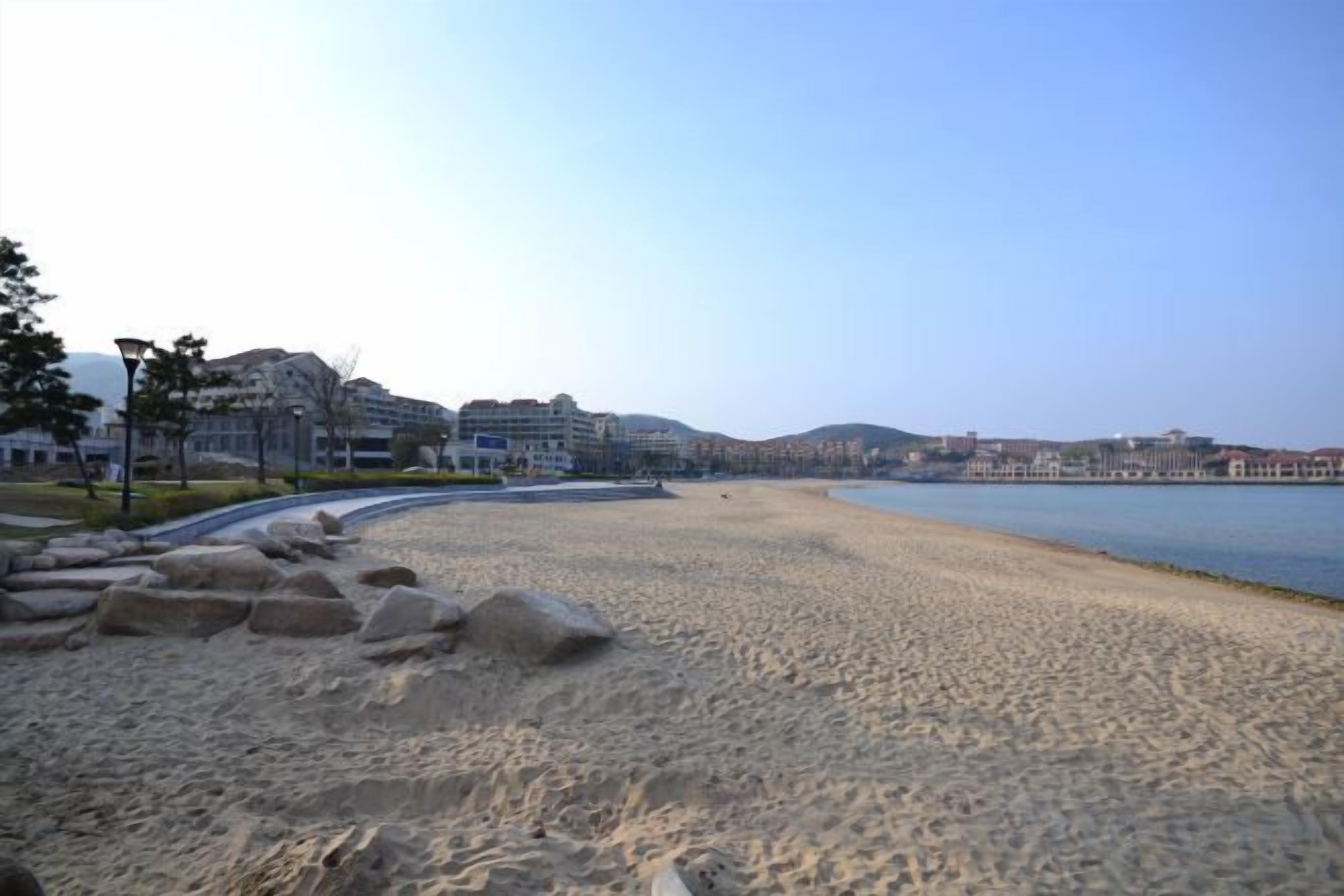 A beach with a clear sky, a sandy shore, and buildings in the background.
