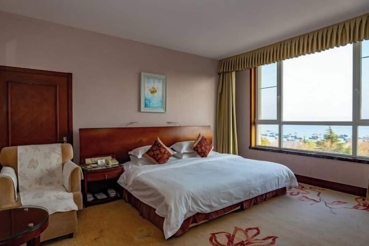 A hotel room with a bed, a chair, a nightstand, a window with curtains, and a painting on the wall.