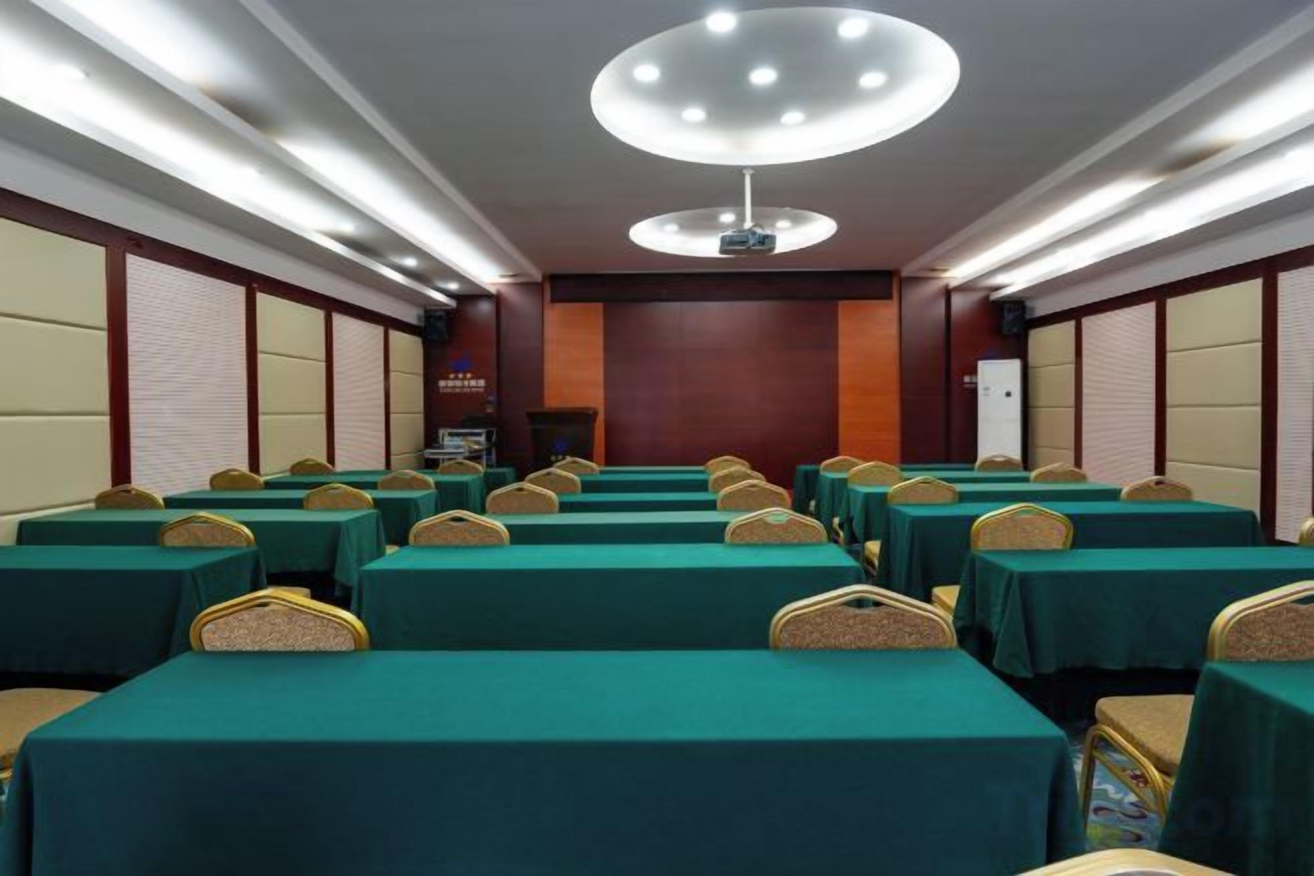 A conference room with rows of tables and chairs, a projector screen, and a large light fixture on the ceiling.