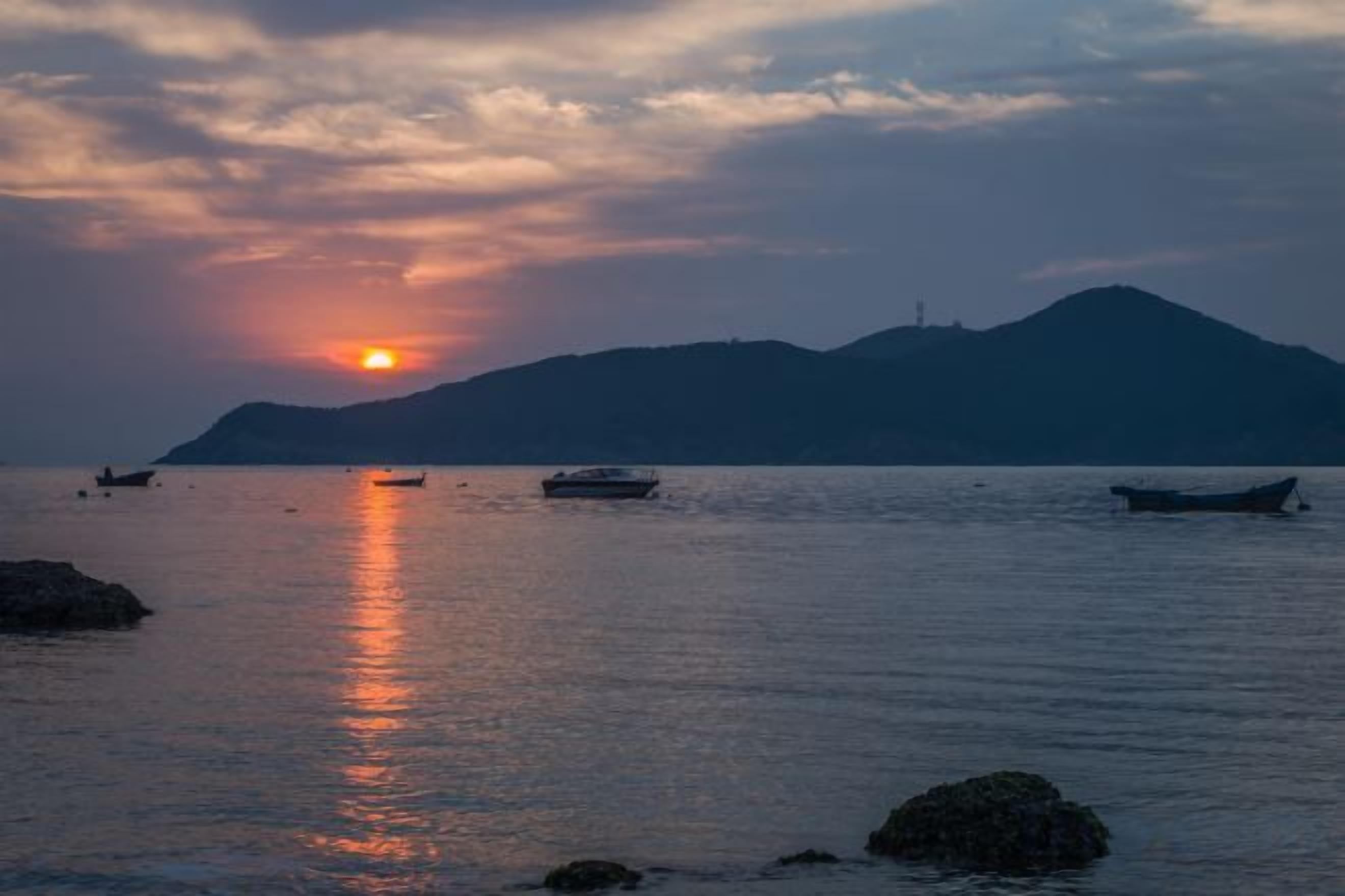A sunset over a calm sea with boats and a mountain in the background.