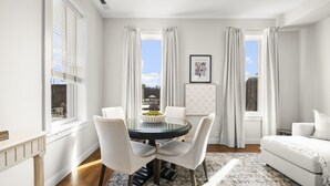 Un salon moderne avec une table ronde en verre, quatre chaises, un canapé et une grande fenêtre avec des rideaux.