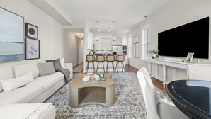 Un salon moderne avec un canapé, une table basse et un téléviseur. Il y a également un coin cuisine avec des tabourets de bar et une table à manger.