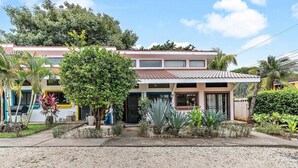 Una casa de dos pisos con techo de tejas, un porche delantero y un pequeño jardín con diversas plantas y un árbol.