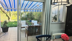 A glass-enclosed patio with a blue and white striped awning, outdoor seating, and a view of a garden.