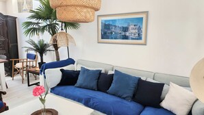 A modern living room with a grey sofa, blue cushions, a wicker lamp, a framed cityscape picture, and a dining area with a wooden table and chairs.
