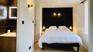 A modern bedroom with a large bed, bedside lamps, a dark headboard, and a bathroom with a mirror and sink. - Charming flat in the heart of Gérardmer (Gérardmer)