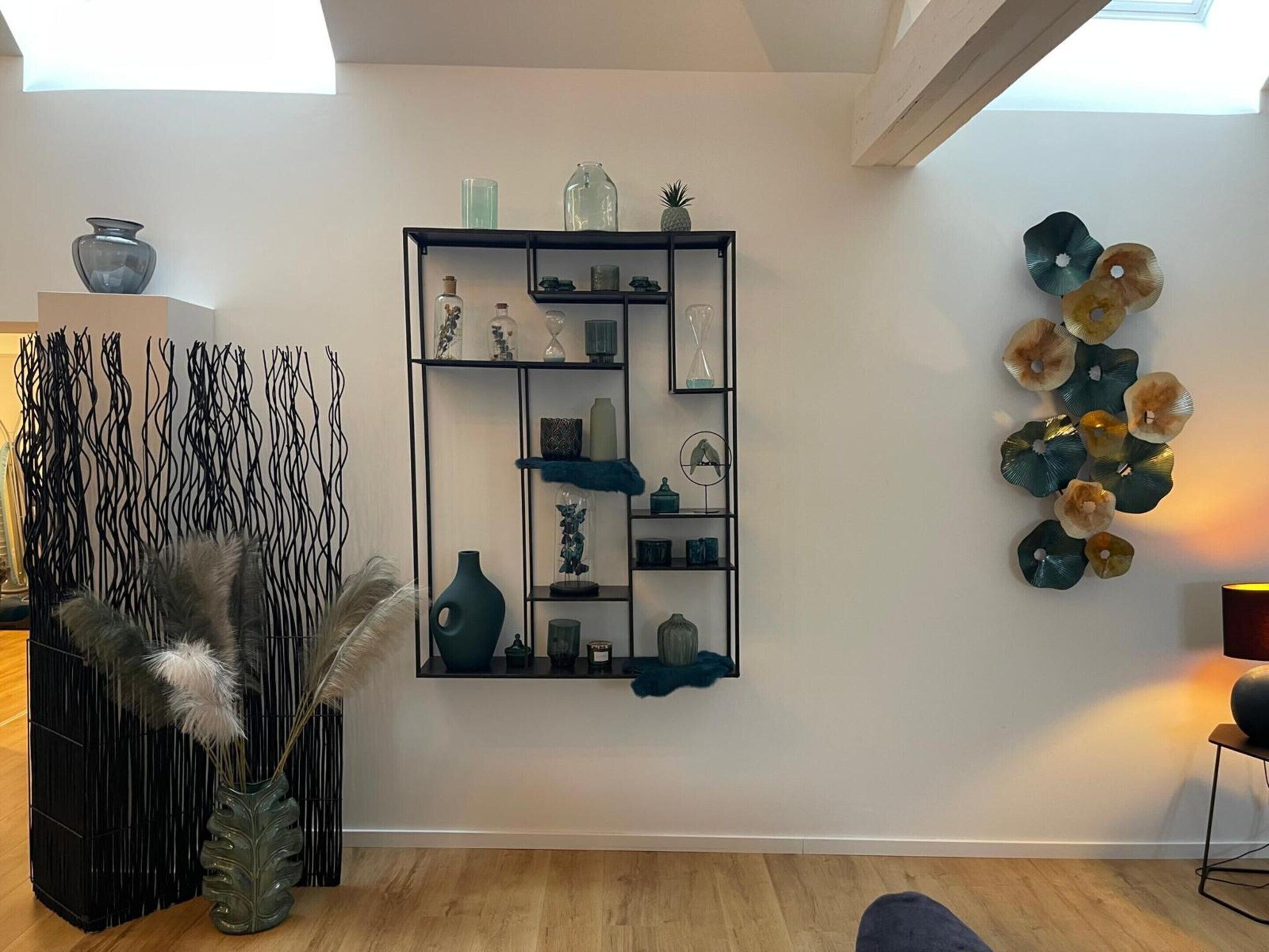 Un interior moderno con una estantería negra que exhibe varios objetos decorativos, una obra de arte colgada en la pared y un arreglo de plantas decorativas.