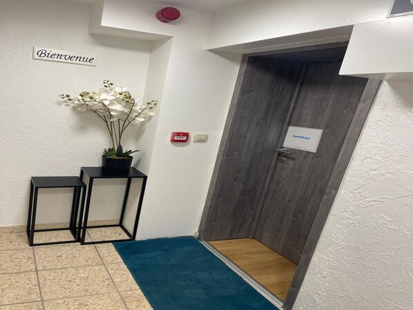 A reception area with a 'Bienvenue' sign, a potted plant, a black metal desk, a blue mat, and a door labeled 'ROOFTOP BLUE'.